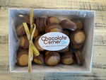 A top-down view of a white gift box filled with chocolate-covered caramel candies, tied with a gold ribbon and featuring a white label that reads 'Chocolate Corner'.